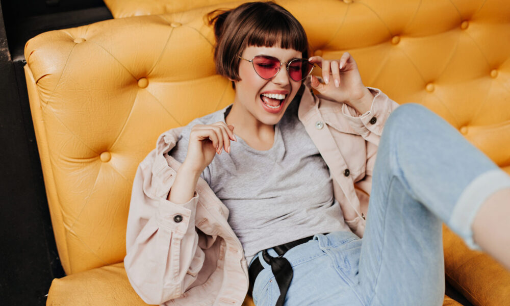 Positive short-haired lady posing on yellow couch in cafe. Brunette girl in jeans, beige jacket and