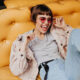 Positive short-haired lady posing on yellow couch in cafe. Brunette girl in jeans, beige jacket and