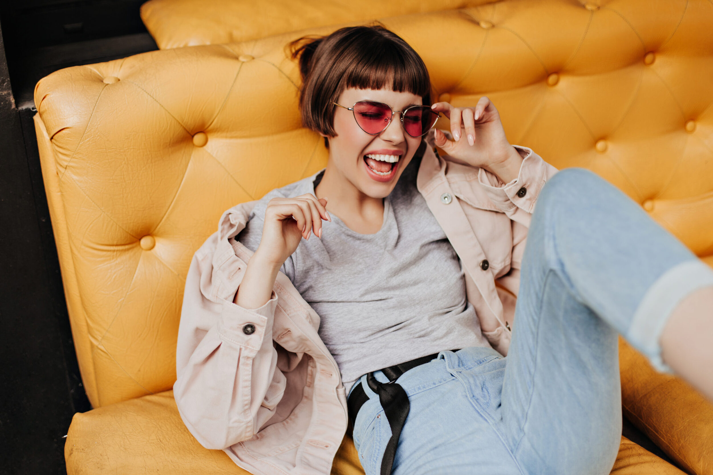 Positive short-haired lady posing on yellow couch in cafe. Brunette girl in jeans, beige jacket and
